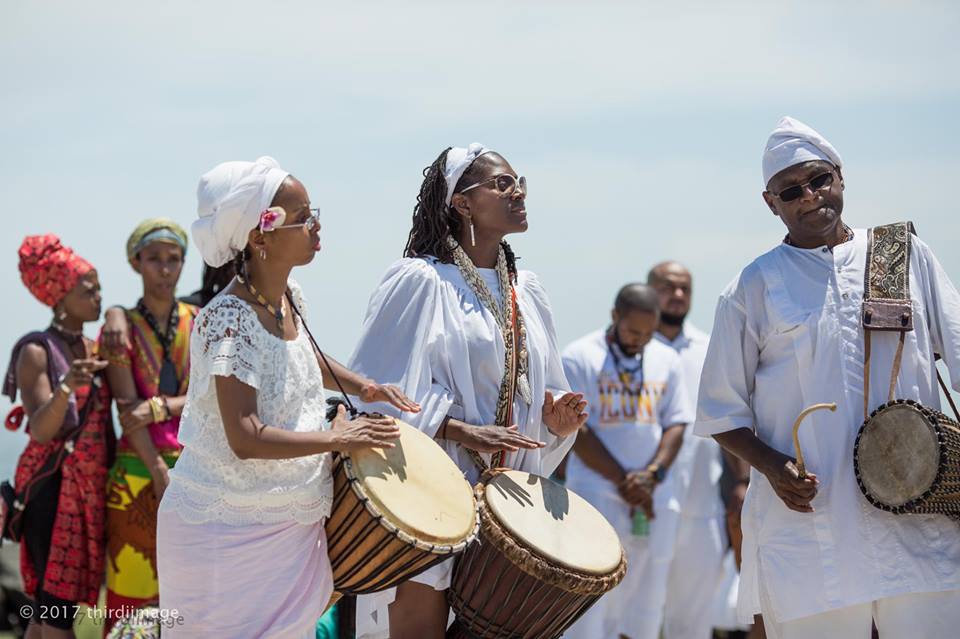Men and women dressed in white and African attire drumming.